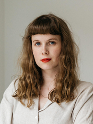 Dara Brexendorf steht vor einer hellen Wand und schaut in die Kamera. Sie trägt ein cremefarbene Bluse und die mittellangen Haare offen.