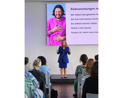 Frau Bössen steht vor Publikum in einem blauen Kostüm und spricht. Hinter ihr eine Leinwand mit Präsentation.