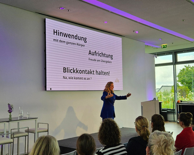 Anna Magdalena Bössen steht vor einem Publikum und spricht mit ausgebreiteten Armen. Sie trägte ein blaues Kostüm und steht vor einer Leinwand, auf der eine Präsentation zu sehen ist.