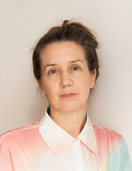 Svenja Leiber lehnt weißen Wand und blickt in die Kamera. Sie trägt eine helle, bunte Bluse und die Haare sind nach hinten zusammengebunden.